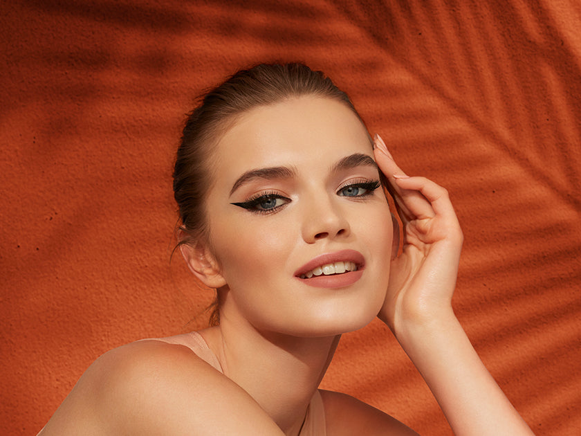 Model with bold winged eyeliner and a flawless makeup look, posing against a warm orange textured background.