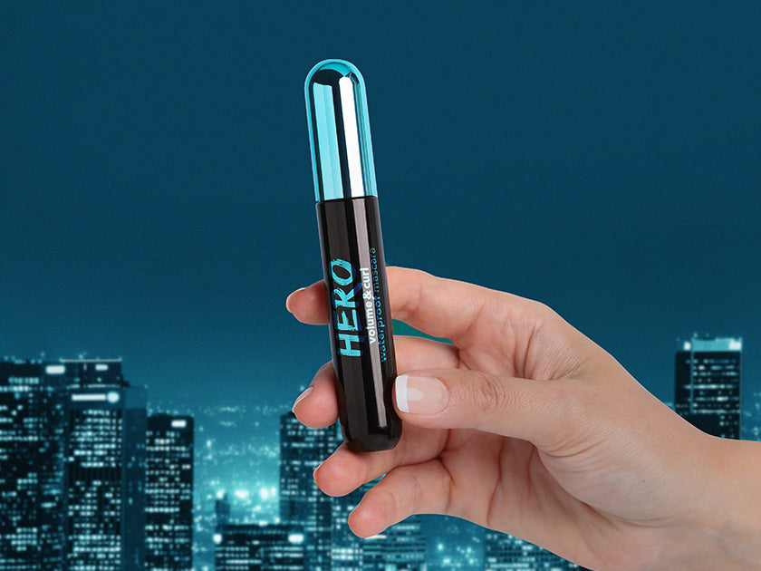 A hand holding Flormar Hero Waterproof Mascara against a nighttime cityscape. The mascara features a sleek black and blue design, emphasizing volume and curl for bold lashes.